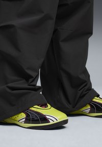 Calzature con un design elegante e curvilineo, caratterizzate da colori giallo neon e nero, con riflessi rosa e una superficie texture. Abbinate a pantaloni neri larghi.