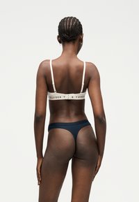 Femme aux cheveux tressés vue de dos, portant un soutien-gorge blanc avec un texte de logo et un string bleu foncé sur un fond clair uni.