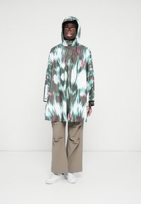 Multicolored poncho in green, gray, and pink with a hood, featuring a tie-dye pattern and loose fit. Paired with beige pants and white sneakers.