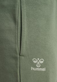 Grüne Baumwoll-Sweatpants mit einer Seitentasche und einem weißen Hummel-Logo am unteren Bein, glatte Textur und lockere Passform.