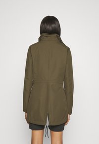 Woman wearing an olive green long-sleeve jacket with a high collar, back slit, and adjustable drawstring visible, paired with black shorts.