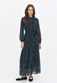 Maxi dress in navy with a floral print, sheer fabric, long sleeves, gathered waist, and ruffle detailing at the hem. Worn with flat shoes.