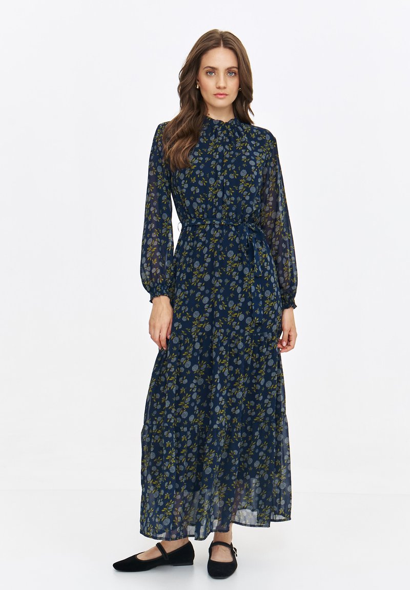 Maxi dress in navy with a floral print, sheer fabric, long sleeves, gathered waist, and ruffle detailing at the hem. Worn with flat shoes.