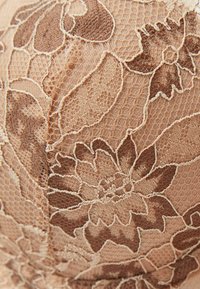Spetslingeri i varm beige med intrikata blommönster och texturerade accenter. Har finkänsliga detaljer och en slät yta.