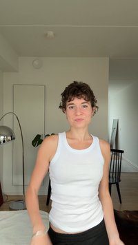 Giovane con capelli ricci corti che indossa una canotta bianca, in piedi in un soggiorno luminoso e minimalista con una lampada da terra e una sedia.