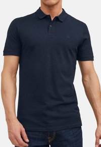 Navyblaue Baumwollpolo mit Kragen, kurzen Ärmeln, zwei Knöpfen und einem kleinen Logo auf der Brust. Glatte Textur.