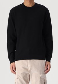 Mann in einem schwarzen Langarmcrewneck-Pullover und beigen Kordhosen, der vor einem schlichten Hintergrund steht.