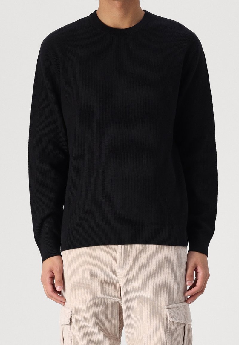 Mann in einem schwarzen Langarmcrewneck-Pullover und beigen Kordhosen, der vor einem schlichten Hintergrund steht.