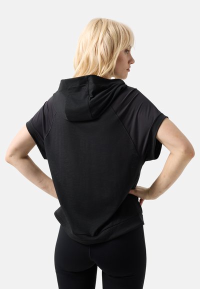 Femme blonde, mains sur les hanches, portant un sweat à capuche noir à manches courtes et des leggings noirs, de dos sur un fond uni.