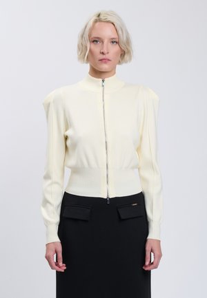 Donna bionda che indossa un maglione chiaro con zip e spalle a sbuffo e una gonna nera con tasche frontali con patta.