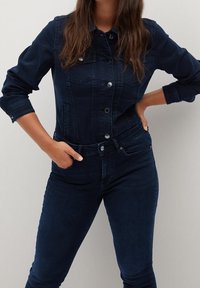 Mujer con chaqueta de mezclilla azul oscuro y jeans a juego, de pie con una mano en la cadera y la otra en el bolsillo contra un fondo liso.