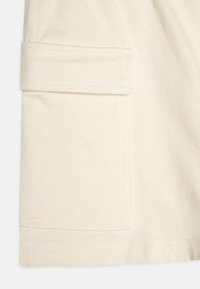 Tejido de algodón de color crema claro que cuenta con un bolsillo lateral con solapa. Textura suave con bordes rectos y costuras bien terminadas a lo largo de las costuras.
