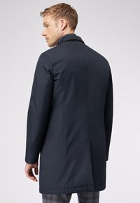 Cappotto blu navy, lungo fino al ginocchio, con una finitura liscia e testurizzata. Presenta un colletto alto, una fessura singola sul retro e un branding discreto.