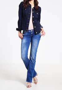 Veste en denim foncé avec jean bleu ; la veste comporte une fermeture boutonnée à l'avant et des poches poitrine ; le jean est légèrement délavé avec une coupe droite.