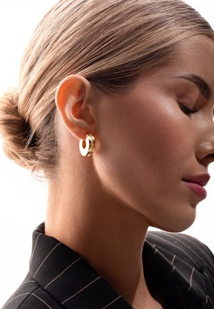 Złote kolczyki w kształcie obręczy mają teksturowaną powierzchnię z polerowanym wykończeniem. Noszone przez osobę w marynarce w paski, na jasnym tle.