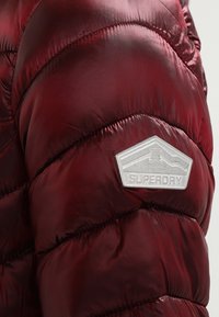 Burgundy pufferjakke lavet af stof med en blank finish, og har et sølvfarvet sekskantet mærke med påskriften "SUPERDRY," fremhævet med stitched sektioner.