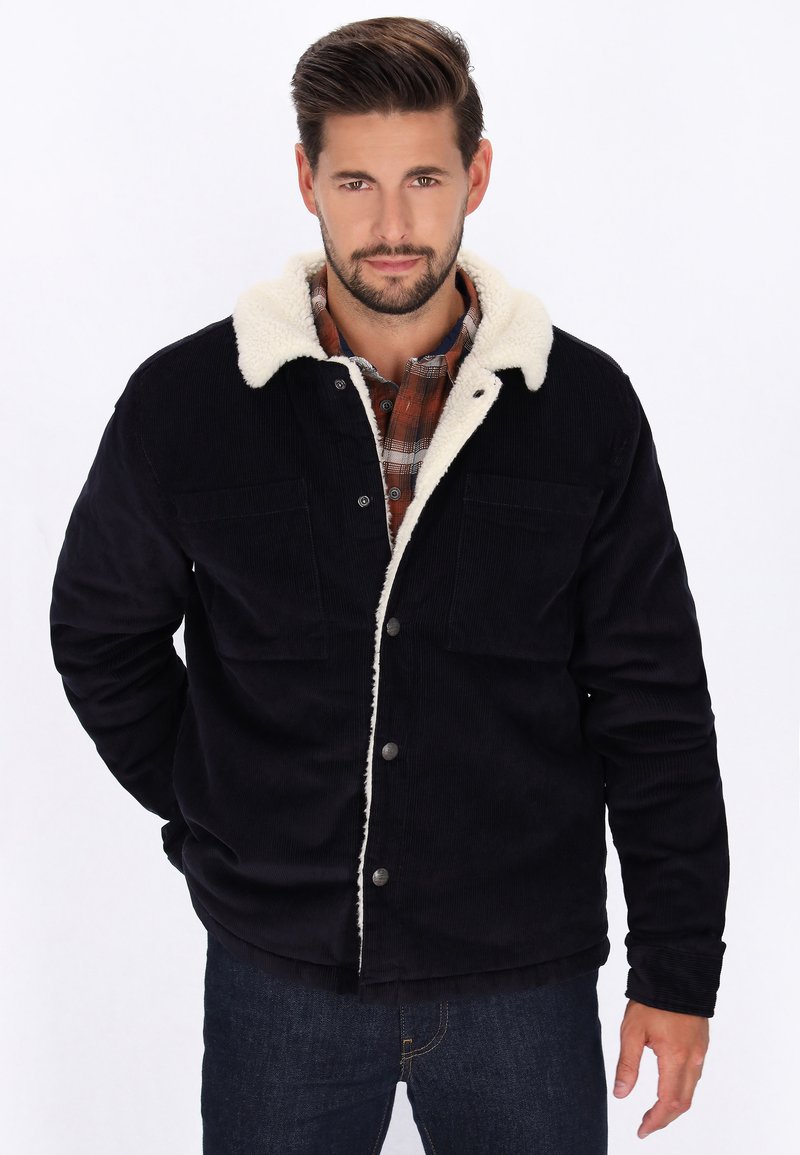 Marineblaue Cordjacke mit einem cremefarbenen Sherpa-Kragen, ausgestattet mit zwei Brusttaschen, Druckknöpfen und einem lockeren Schnitt. Über einem karrierten Hemd getragen.