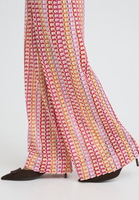 Femme portant un pantalon large à motifs rose, orange et rouge, avec des chaussures marron à talons pointues, sur un fond blanc.