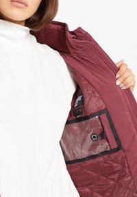 Chaqueta acolchada burdeos con interior acolchado, que incluye un bolsillo transparente y un botón de control. Tejido liso, cuello de punto visible.