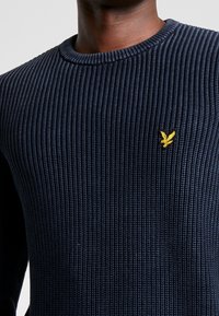 Marineblauer gerippter Strickpullover mit Rundhals, ausgestattet mit einem kleinen gelben Emblem auf der Brust. Die Textur ist deutlich erhaben und strukturiert.