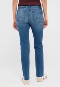 Une personne portant un jeans droit bleu moyen et des baskets blanches, se tenant de dos face à l'objectif sur un fond clair.