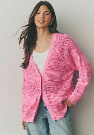 Cardigan rose léger en maille avec un motif texturé, décolleté en v et deux poches avant, associé à un débardeur blanc et à un jean bleu.