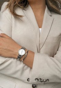 Silver wristwatch with a white face, worn on a wrist with a silver bangle. Beige suit jacket with a subtle pocket and button detail.