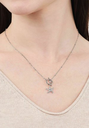 Close-up van een persoon die een zilveren ketting met een toggle-sluiting en een zeestervormige hanger draagt.