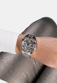 Srebrna ročna ura z okroglo površino, kristalnimi akcenti in večimi številčnicami. Ima kovinski pas in se nosi na zapestju z teksturirano manšeto.