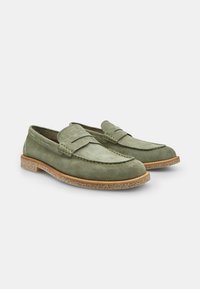Mocasines de ante verde con punta redondeada, detalles de costura y una suela texturizada marrón. Diseño casual con un estilo cómodo para poner y quitar fácilmente.