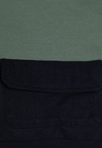 Grønt bomullsoberste med en mørk denim underdel som har en stor lommeklaff. Glatt tekstur med kontrast mellom fargene.