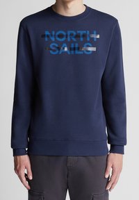 Muž nosící námořnickou modrou mikinu s nápisem "NORTH SAILS" v modré, překřížený dvěma horizontálními stříbrnými a černými tahy štětce.