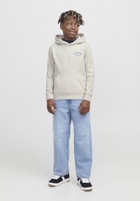 Felpa color beige chiaro con tasca a marsupio e testo blu, abbinata a jeans azzurri e sneaker nere con accenti bianchi.