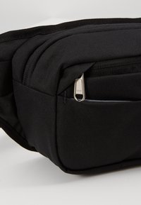 Sac banane en tissu noir avec une poche avant zippée équipée d'une tirette de fermeture éclair YKK argentée.