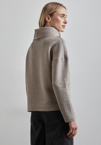 Vaalean beige ribattu neule, jossa on korkea kaulus, laskeutuvat olkapäät ja pehmeä rakenne, yhdistettynä tummiin farkkuihin. Taustalla on neutraali harmaa.