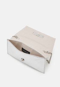 Petit sac à main argenté avec bandoulière chaîne, rabat ouvert montrant l'intérieur beige et l'étiquette de la marque à l'intérieur.