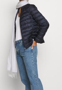 Person wearing navy quilted jacket, white top, blue jeans, white scarf, and gold hoop earring standing with one hand in pocket.