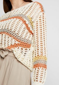 Pull en crochet crème avec des rayures orange, grises et jaunes. Design à tissage ouvert, manches longues et coupe décontractée. Associé à un pantalon beige.