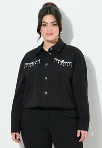 Mustassa lyhyt takissa on nappikiinnitys, etutaskut ja koristeelliset helmikoristeet rinnassa. Kankaan rakenne on teksturoitu ja helma on rispaantunut.