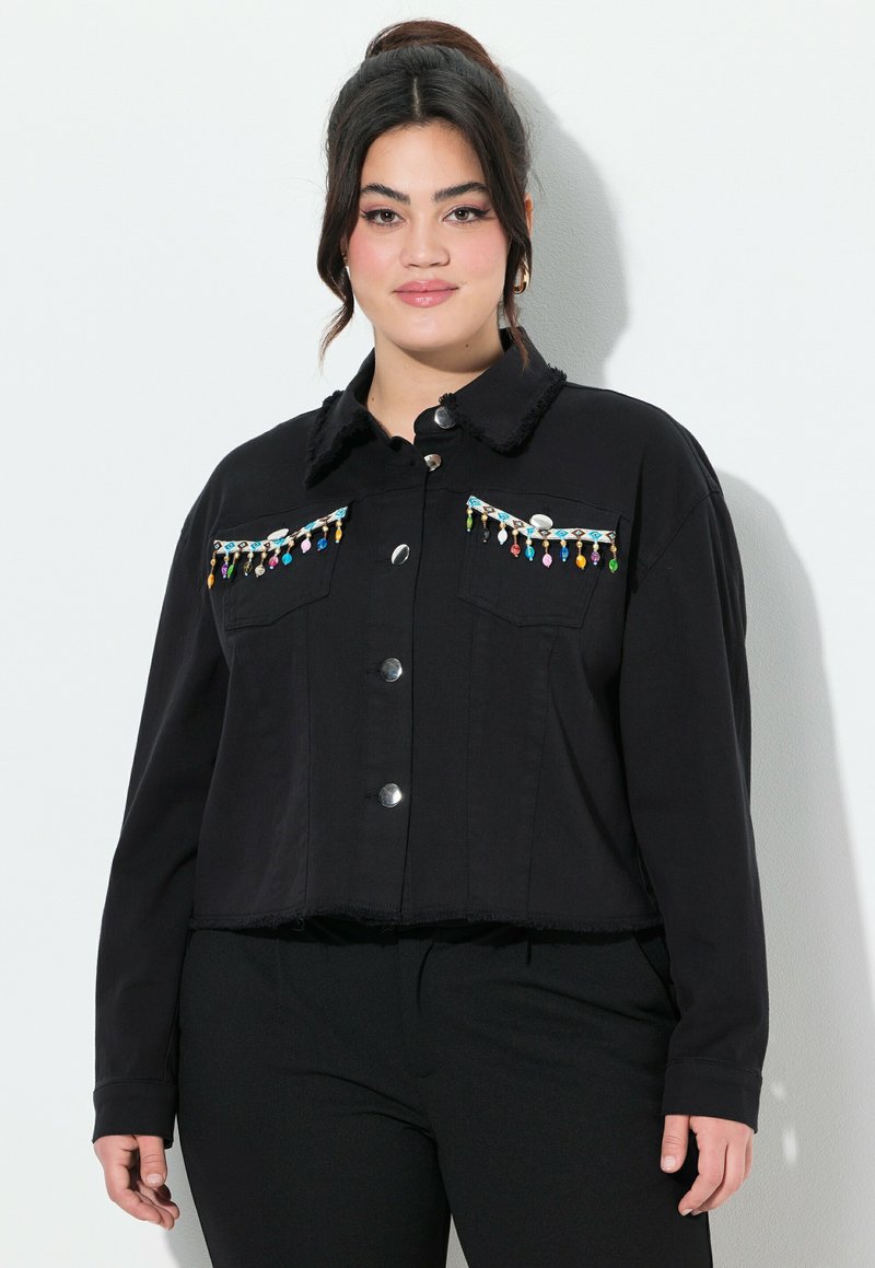 Mustassa lyhyt takissa on nappikiinnitys, etutaskut ja koristeelliset helmikoristeet rinnassa. Kankaan rakenne on teksturoitu ja helma on rispaantunut.