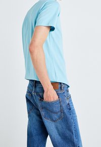 Ljusblå bomull t-shirt med kort ärm parat med blå denim jeans som har en bakficka och en brun läderlapp på midjebandet.