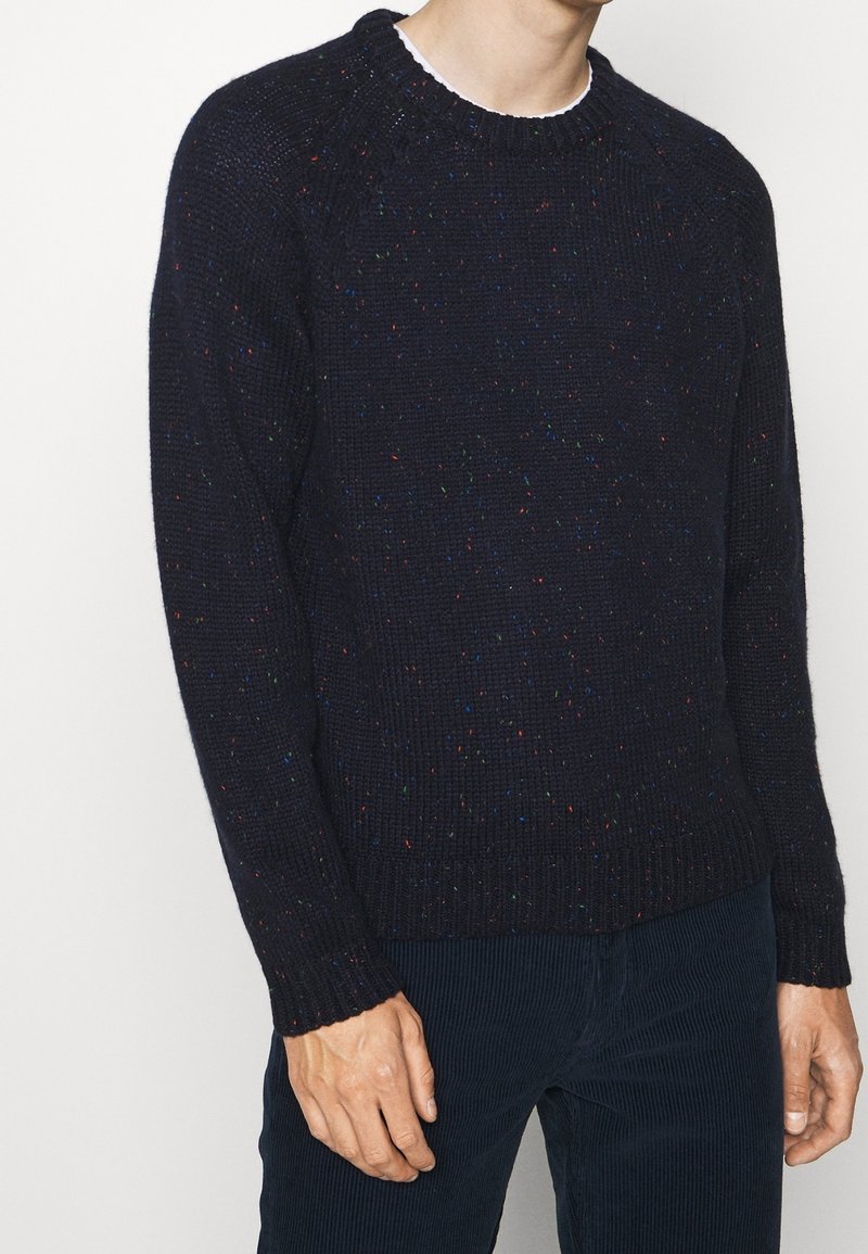 Homme portant un pull en maille bleu marine foncé moucheté par-dessus une chemise blanche et un pantalon en velours côtelé foncé, debout devant un fond uni.