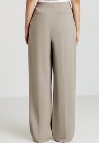 Femme portant un pantalon beige à taille haute et jambes larges avec des poches arrière passepoilées, se tenant devant un fond léger uni.