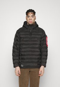 Felpa nera imbottita con design a righe orizzontali, chiusura zip sul davanti, cappuccio regolabile e accento rosso sulla manica. Indossata con pantaloni marroni.