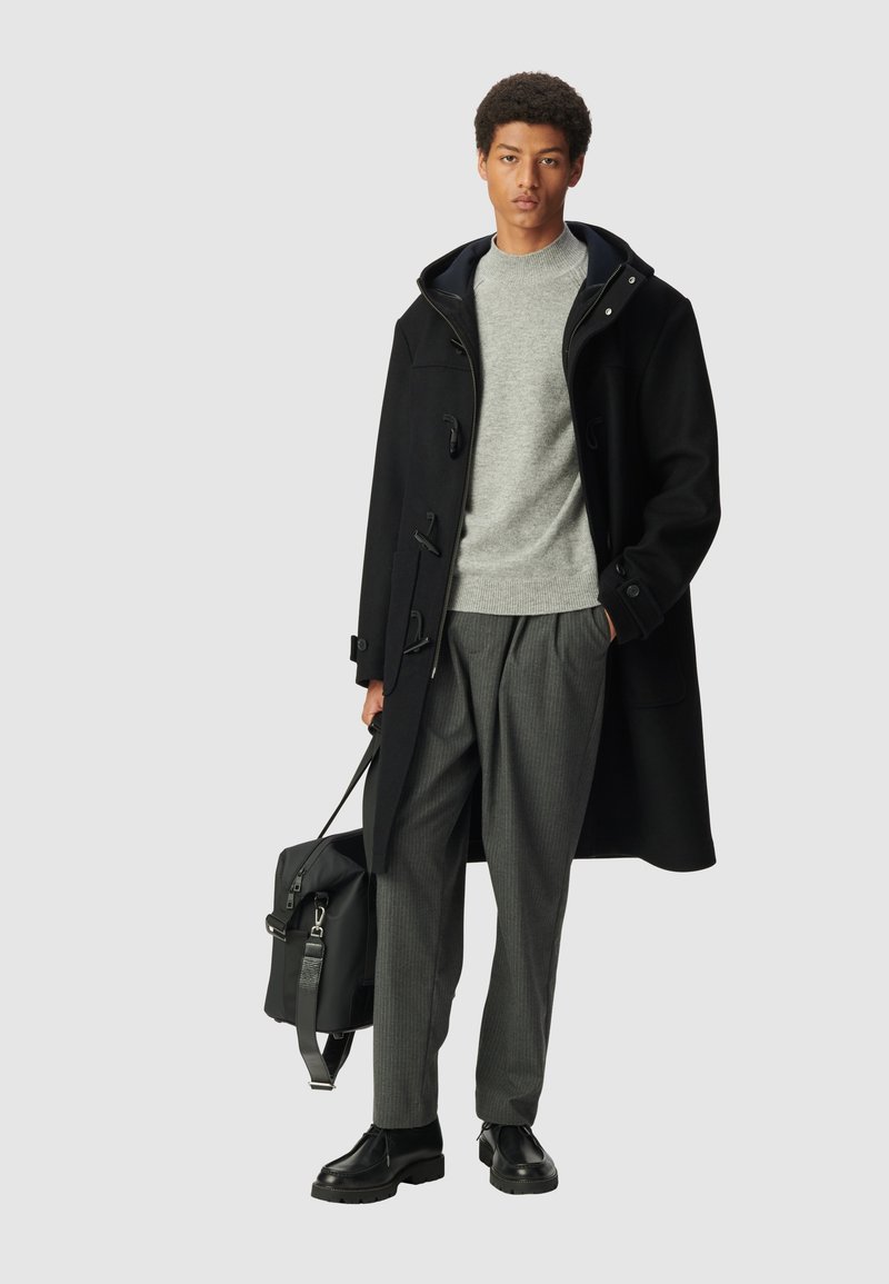 Cappotto duffle nero con chiusure a un pulsante, maglione grigio a maglia, pantaloni a righe e scarpe in pelle nere. Borsa nera con tracolla.