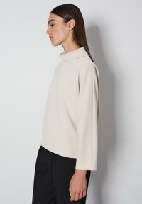 Beige lange mouw top met een hoge halslijn en details aan de zijnaad, gemaakt van gladde stof. Gepaard met zwarte broeken.