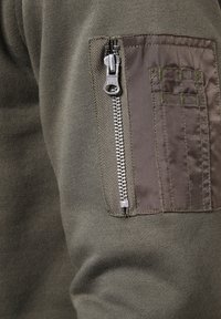 Olijfgroene sweatshirtstof met een ritssluiting zak en een donkergrijze paneel. Inclusief zilveren ritsbeslag en subtiele stikdetails.