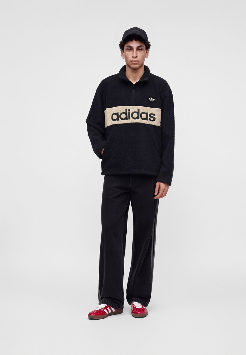 Νέος άνδρας στέκεται φορώντας μαύρο fleece Adidas με μπεζ λογότυπο, μαύρο παντελόνι, μαύρο καπέλο και κόκκινα αθλητικά παπούτσια Adidas με λευκές ρίγες.