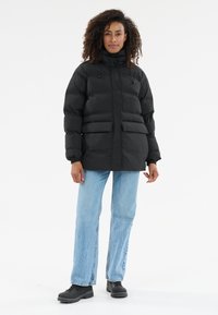 Kvinde står i en sort dunjakke, lyseblå jeans og sorte støvler foran en hvid baggrund.