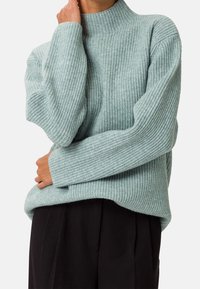 Jasnoniebieski sweter z prążkowanego dzianiny z wysokim kołnierzem, długimi rękawami i luźnym krojem. Teksturowany materiał oraz opadające ramiona, tworzące swobodną sylwetkę.